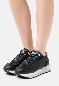 Black leather sneakers with white and mint green accents, featuring branded laces, a thick sole, and a minimalist design.