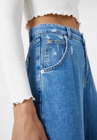 Zbliżenie na osobę w wysokich, poszarpanych, niebieskich dżinsach oraz białym, prążkowanym, krótkim topie z długim rękawem i falbanowymi brzegami.