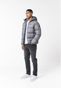 Chaqueta de plumón gris con capucha, con cierre frontal y puños elásticos, llevada sobre una camiseta blanca, jeans gris oscuro y zapatillas blancas.