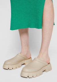 Stopy w beżowych, masywnych butach wsuwanych, zestawione z zieloną, prążkowaną spódnicą, na jednolitym jasnym tle.