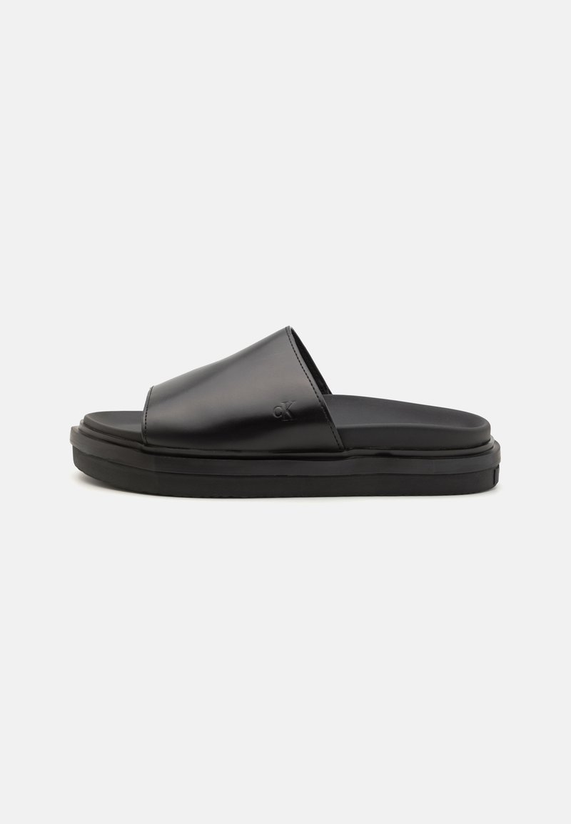 Chinelas pretas de slip-on com um cabedal de couro liso e sola de borracha espessa. O design é minimalista, sem enfeites visíveis.