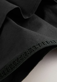 Gros plan sur un tissu noir avec une bordure brodée comportant le texte répété "BAKER" le long du bord.