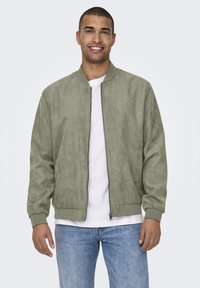 Olivgrüne Bomberjacke aus weichem, wildlederähnlichem Material mit einem Reißverschluss vorne, gerippten Bündchen und Saum. Getragen über einem weißen T-Shirt und Jeans.