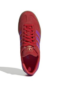 Czerwona zamszowa sneaker z fioletowymi paskami, złotym logo, teksturowaną wkładką i gumową podeszwą. Wierzch posiada klasyczne zapięcie na sznurówki.