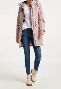 Ljusrosa regnjacka med huva, framsidans dragkedja, elastiska ärmslut och justerbara snoddar. Sammanvävd med blå jeans och beige snörstövlar.