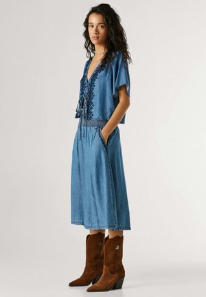 Femme portant un haut en denim bleu brodé avec une jupe midi assortie et des bottes cow-boy en daim marron mi-mollet, debout les mains dans les poches.