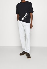 Černé tričko s krátkým rukávem a bílým potiskem "Yrtrn", spárováno s volnějšími světle modrými džíny a hnědými šněrovacími botami.