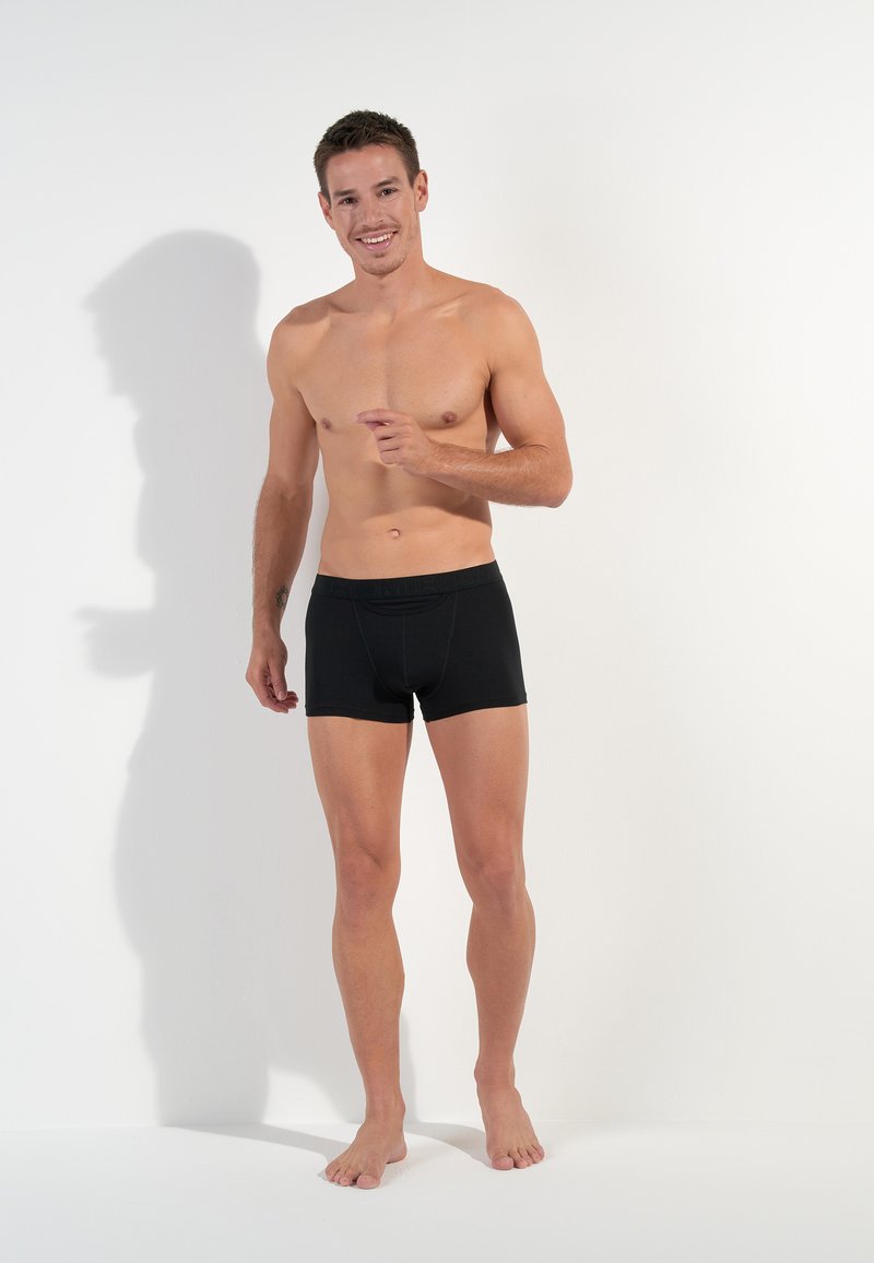 Schwarze Boxershorts aus dehnbarem Stoff, mit einem bequemen Bund. Das Modell steht barfuß vor einem schlichten weißen Hintergrund.