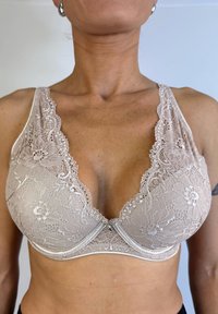 Bralette de encaje en beige con escote en V profundo, patrones florales y detalles decorativos. Cuenta con copas suaves y tirantes ajustables.