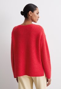 Cozy knit sweater in bright red, featuring a loose fit, wide sleeves, and a ribbed texture. Designed with a rounded neckline and side slits.