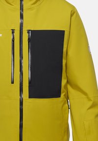 Gelbe Jacke mit schwarzem Brusttasche, Reißverschluss, langen Ärmeln und strukturiertem Stoff. Verfügt über schwarze Reißverschlusszüge und ein Logo auf dem Ärmel.