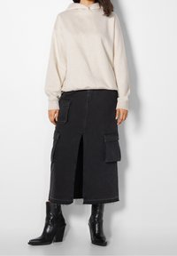 Beige huvtröja, svart lång denimkjol med frontrisk och dubbla cargo-fickor, tillsammans med svarta klackade ankelstövlar.