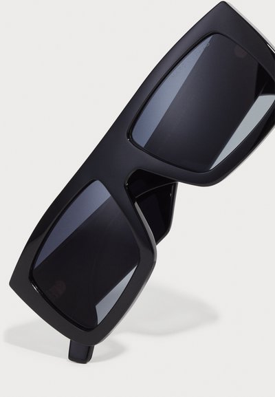 Schwarze übergroße Sonnenbrille mit mattem Finish, eckigen Rahmen und dunklen Gläsern. Verfügt über eine auffällige Markengravur an der Seite.
