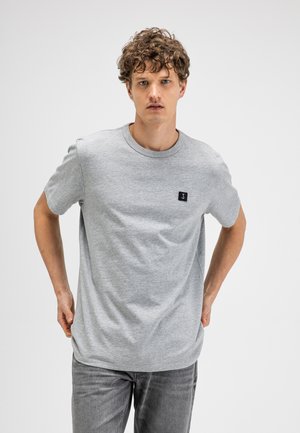 Jonge man met krullend haar, die een effen lichtgrijs T-shirt en grijze spijkerbroek draagt, staand tegen een witte achtergrond.