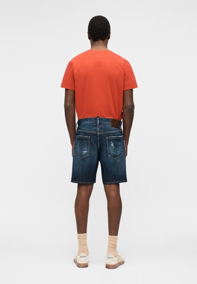 Άνδρας που φοράει πορτοκαλί μπλουζάκι, μπλε τζιν σορτς με πίσω τσέπες, μπεζ κάλτσες και μπεζ αθλητικά παπούτσια, στέκεται με την πλάτη προς τα εμπρός σε μονόχρωμο φόντο.