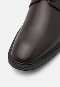 Chaussure en cuir marron avec une finition polie, bout rond et texture subtile. Dispose d'une semelle noire à profil bas et de coutures décoratives.