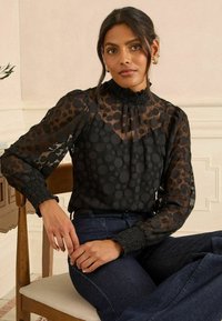 Crna bluzica s točkicama od prozirnog materijala, visoki naberani ovratnik, puf rukavi i smockani manžeti. U kombinaciji s tamnim trapericama.