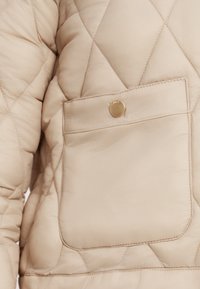 Πουπουλένιο μπουφάν ανοιχτό μπεζ με μεγάλο τσεπάκι με καπάκι και κλείσιμο με χρυσό κουμπί και αντιθετικές ραφές. Μαλακή, επενδεδυμένη υφή.