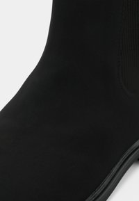 Stivale alla caviglia in suede nero con una texture liscia, pannelli laterali elasticizzati e punta arrotondata. La suola è piatta con una leggera lucentezza.