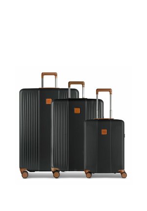 Trois valises rigides noires avec des poignées marron et quatre roues chacune, disposées en rangée de la plus grande à la plus petite.