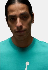 T-shirt in cotone teal con maniche corte e colletto tondo, con un logo bianco al centro. Tessuto morbido e design informale.