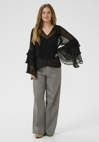 Top nero trasparente a maniche lunghe con volant sovrapposti, abbinato a pantaloni a gamba larga grigi con motivo a scacchi e scarpe beige.