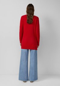 Kobieta z długimi brązowymi włosami, odwrócona tyłem, ubrana w długi czerwony sweter i szerokie jasnoniebieskie dżinsy, na jednolitym szarym tle.