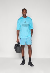 Šviesiai mėlyna „Adidas“ sportinė marškinėliai ir šortai su juodu logotipu, derinami su juodais sportiniais batais ir tamsia sportine kuprine.