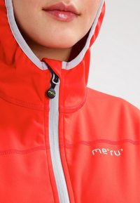 Chaqueta impermeable roja con un alto cuello, cremallera y costuras grises, que presenta el nombre de la marca "me'ru'" en blanco. Textura suave.