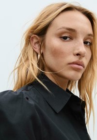 Camicia nera con colletto e maniche a sbuffo, indossata da un modello con lunghi capelli biondi, che mette in evidenza una texture liscia e fine con dettagli minimi.