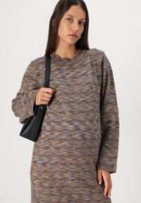 Robe en tricot marron avec des motifs ondulés bleus et beiges. Dotée d'un col rond et de manches longues. Associée à un sac à main noir.