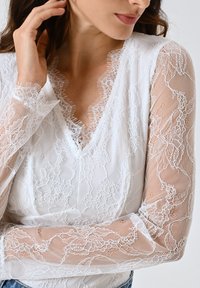 Weißes Bluse mit durchscheinenden Spitzenlangen Ärmeln mit floralen Mustern, einem V-Ausschnitt und gewellten Kanten, die eine zarte, schichtartige Textur schaffen.