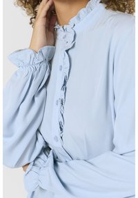 Personne portant un chemisier bleu clair avec patte de boutonnage à volants et poignets froncés, main touchant le cou, cheveux bouclés partiellement visibles.