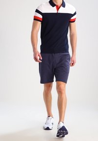Marinfärgad polo med röda, vita och marinblå ränder på axlar och krage, kombinerad med mörkgrå shorts och vita sportskor.