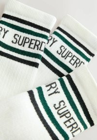 Hvide bomuldsstrømper med to grønne og en sort horisontal stribe, med "SUPERDRY" strikket ind i kanten. Ribbet struktur.