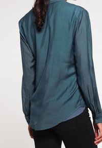 Blusa di seta turchese con piega posteriore, maniche lunghe, polsini con bottone e orlo curvo, dal texture morbido e leggero luccichio.