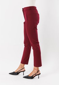 Pantaloni slim-fit color bordeaux con tasche laterali, abbinati a décolleté nere a punta e una cavigliera rossa su uno sfondo chiaro.