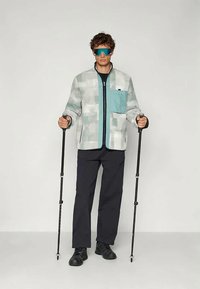 Άνδρας που φοράει μοτίβο σακάκι και γυαλιά ηλίου, κρατώντας μαύρα μπόσικα πεζοπορίας, στέκεται μπροστά σε απλό φωτεινό φόντο.