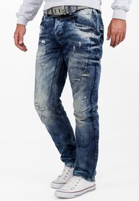 Denim jeans in faded blue with distressing and frayed patches. Slim fit design, featuring five pockets and a gray belt, worn with white sneakers.