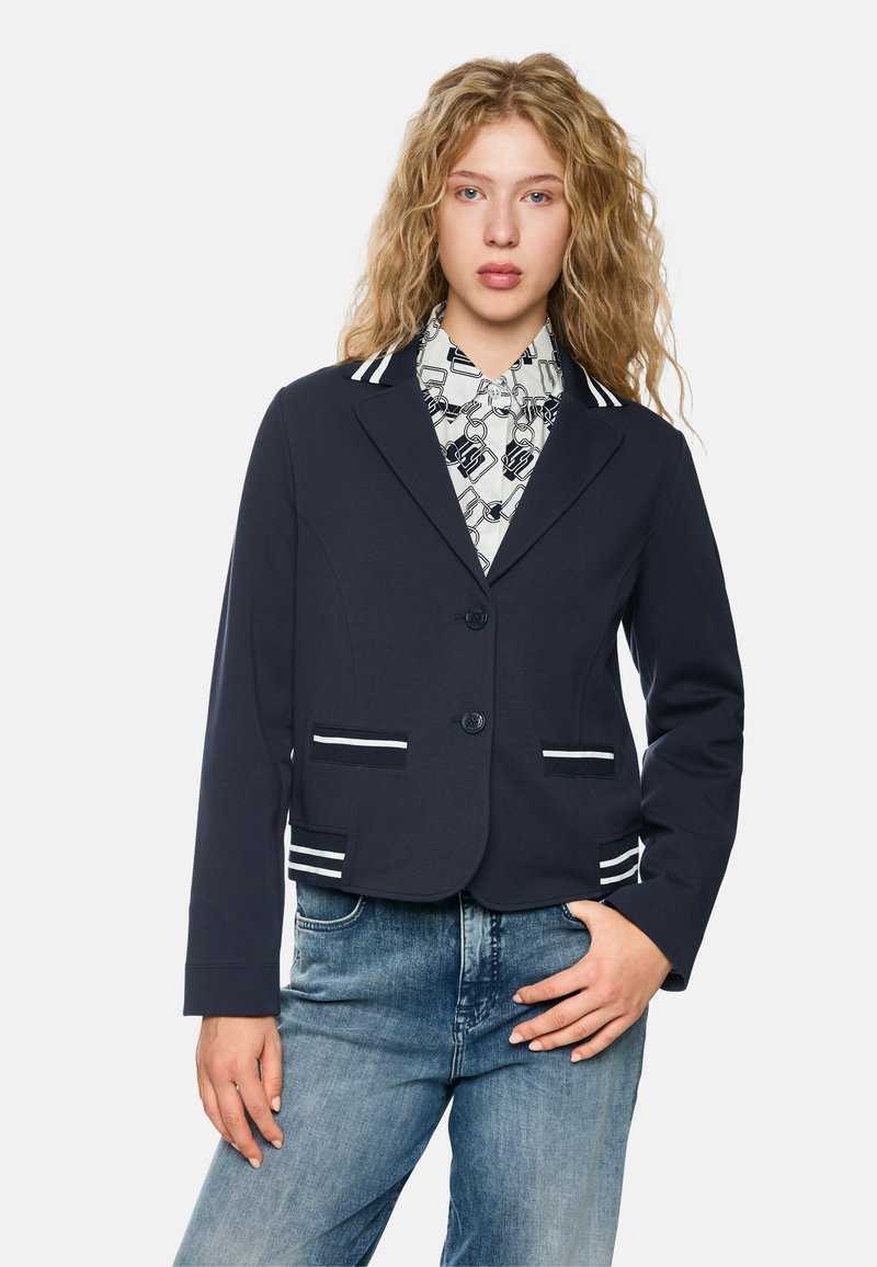 Navy blazer with white accents, two front pockets, single button closure, and a smooth texture. Shirt underneath features a geometric pattern.