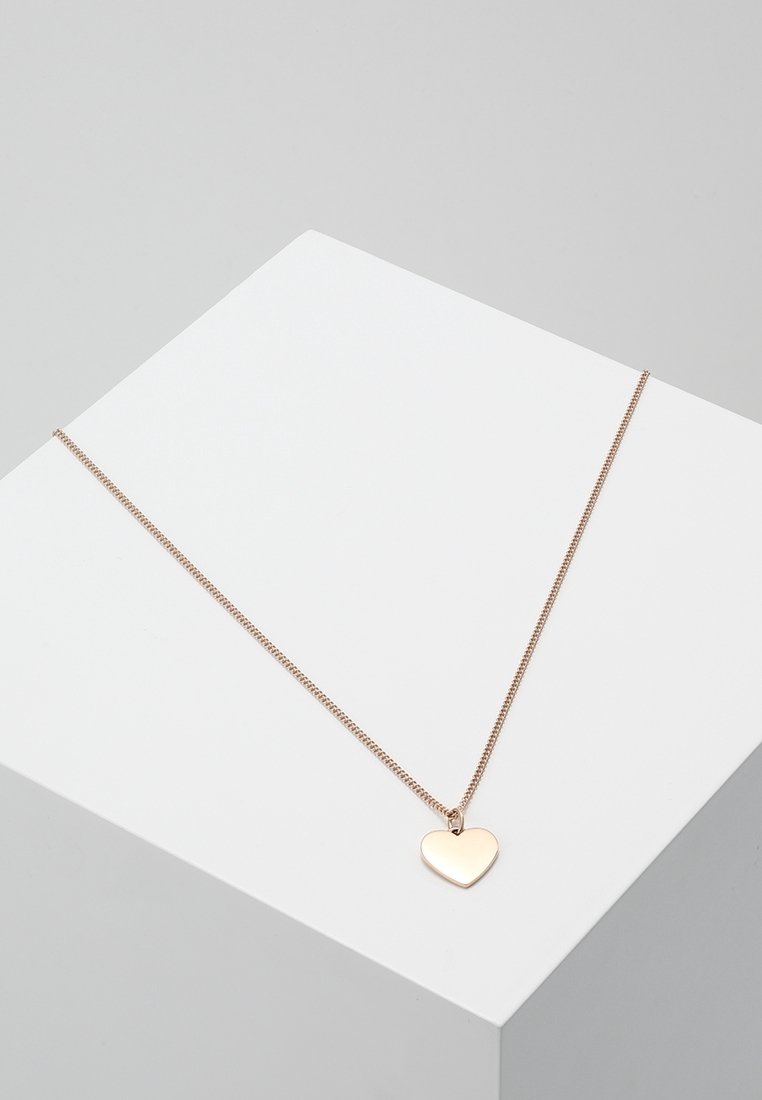 Collier fin en chaîne d’or avec un pendentif lisse et simple en forme de cœur reposant sur un piédestal géométrique blanc sur fond gris.