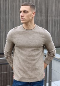 Vaaleanruskea tekstuuri neulepaita pyöreällä pääntiellä, pitkät hihat ja resorit manseteissa, yhdistetty sinisiin farkkuihin.