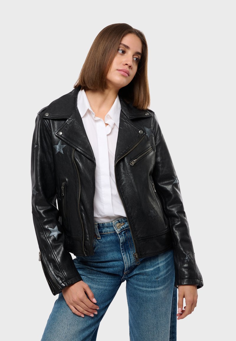 Crna kožna motociklistička jakna sa srebrnim zvjezdastim detaljima, asimetričnim zatvaranjem na zip, i širokim reverima, nosi se preko bijele košulje s gumbima.