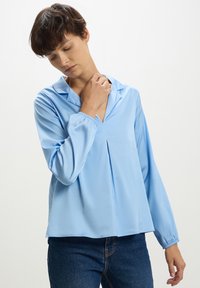 Blusa azzurro chiaro a maniche lunghe in tessuto liscio con scollatura a V e dettagli plissettati sulla parte anteriore. Polsini elasticizzati e vestibilità comoda, abbinata a jeans scuri.
