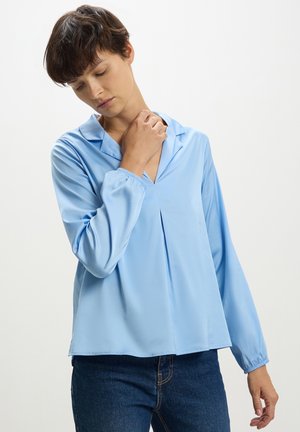 Blusa azzurro chiaro a maniche lunghe in tessuto liscio con scollatura a V e dettagli plissettati sulla parte anteriore. Polsini elasticizzati e vestibilità comoda, abbinata a jeans scuri.