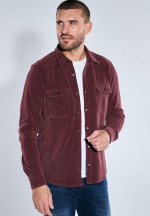 Mann trägt ein weinrotes Cordhemd über einem weißen T-Shirt und blauen Jeans, schaut nach links vor einem einfarbigen Hintergrund.