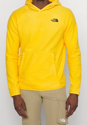 Man draagt een felgele fleece hoodie van The North Face en beige cargobroek, staand met ontspannen handen langs zijn zij.