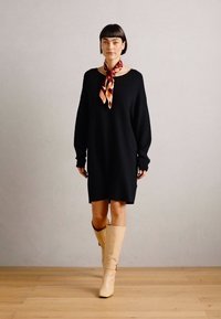Robe-sweater noire oversize à manches longues, associée à une écharpe multicolore et des bottes crème montant jusqu'aux genoux, sur un mannequin se tenant sur un sol en bois.