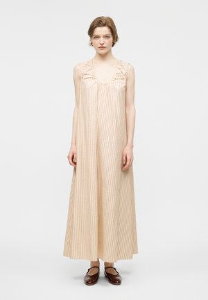 Žena stoji noseći bezrukavnu bež dugu haljinu s tankim okomitim prugama i smeđe Mary Jane cipele na bijeloj pozadini.