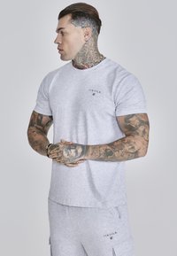 T-shirt gris à manches courtes au style ajusté, avec un petit logo noir sur la poitrine. Fabriqué en tissu doux semblable à du jersey.